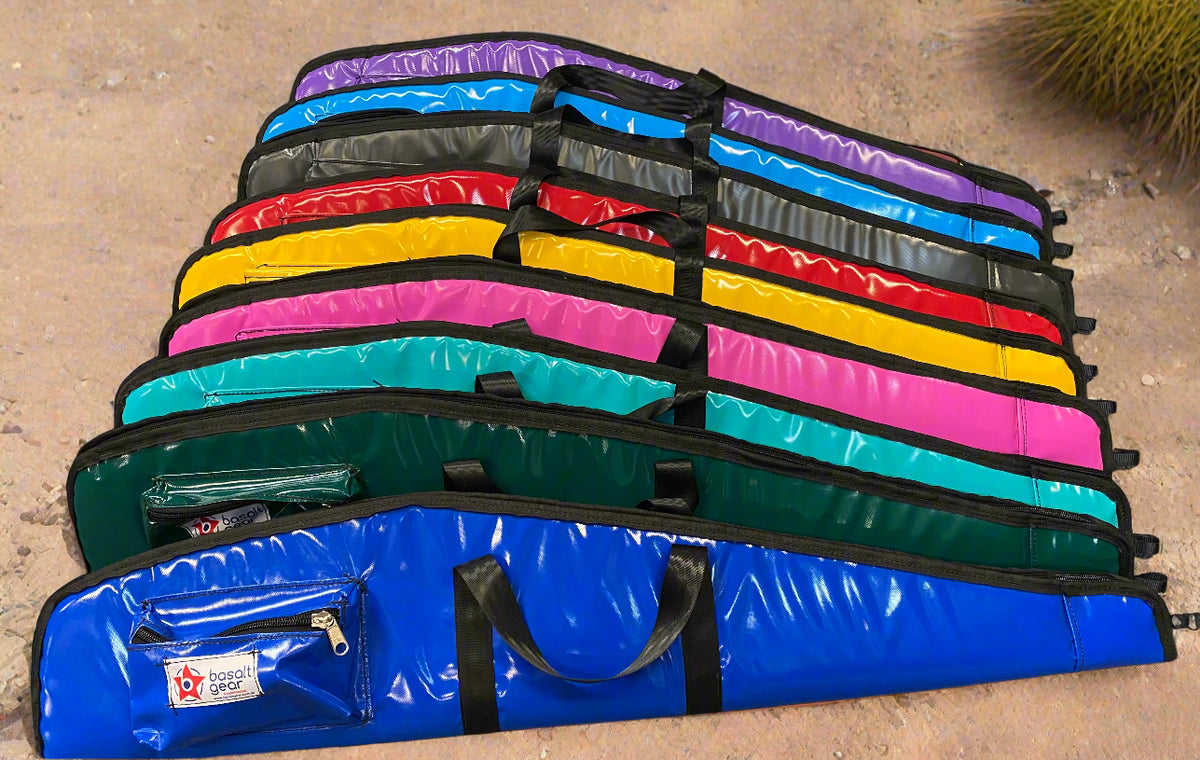 A collection of Basalt Gear vinyl gun bags in various colors, including black, green, pink, yellow, purple, blue, and red. They are laid out on the floor with zippers and carry handles visible.