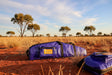 A Royal blue waterproof gear bag with a yellow label lies on red earth in an Australian outback landscape, with another matching bag partially visible in the foreground and trees under a blue sky in the background.